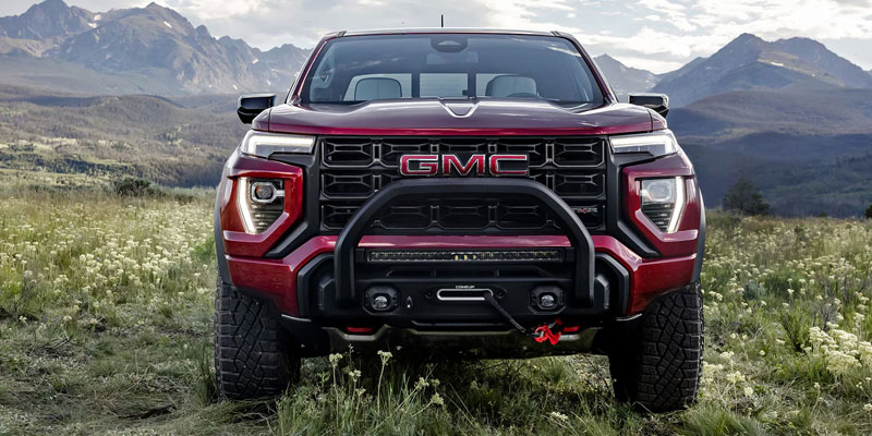 A front view of a GMC truck with a bold grille, off-road tires, and a bumper guard, set against a mountainous landscape.